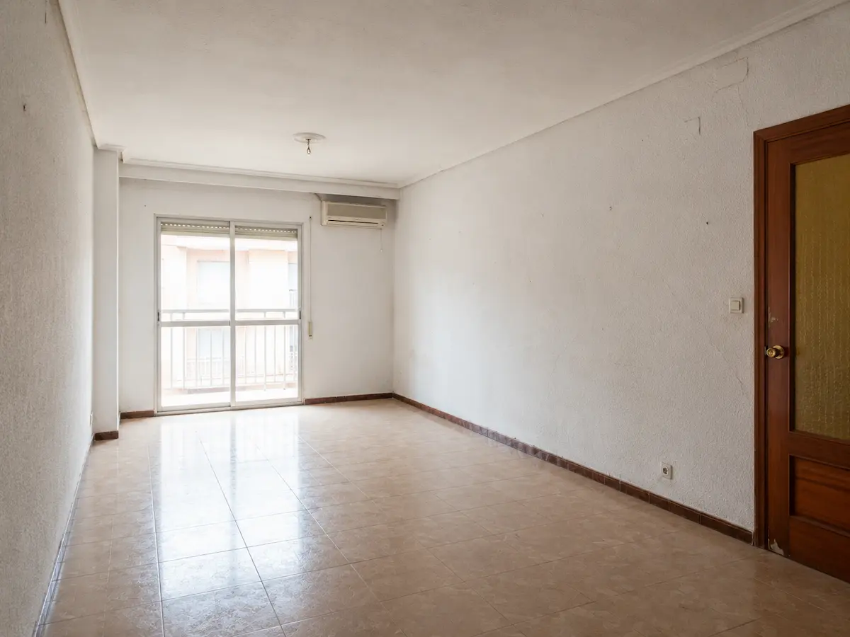 Salón de una vivienda en Valencia antes de la reforma, con paredes de gotelé, grietas, puerta de madera y suelo cerámico antiguo.